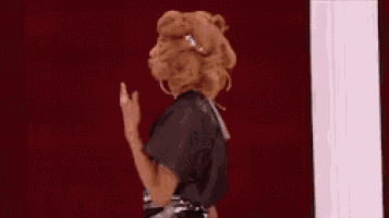 a woman in a wig is standing in front of a red curtain and making a funny face .