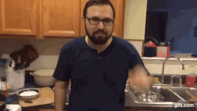 a man with a beard and glasses is standing in front of a sink in a kitchen .