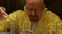 a man with a beard and glasses is sitting at a table with beakers .