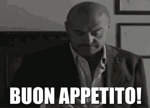 a bald man in a suit is sitting at a table with the words buon appetito written on the screen .