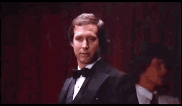 a man in a tuxedo is standing in front of a red curtain smoking a cigarette .