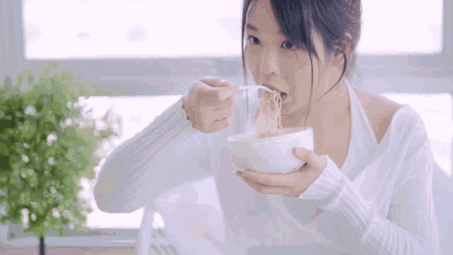 a woman in a white sweater is eating noodles from a white bowl with a spoon