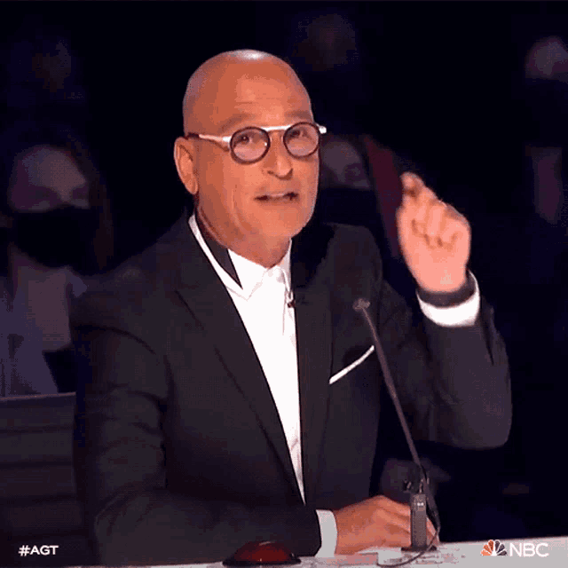 a bald man wearing glasses and a suit is speaking into a microphone with the nbc logo behind him