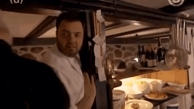 a man in a white shirt is standing in a kitchen with a bottle of wine on a shelf .