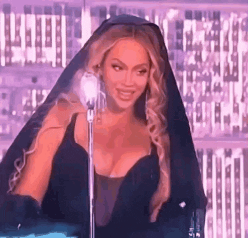 a woman in a black dress and veil is standing in front of a microphone and smiling .