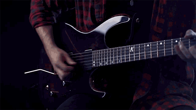 a man in a plaid shirt is playing a guitar with the letter k on the neck