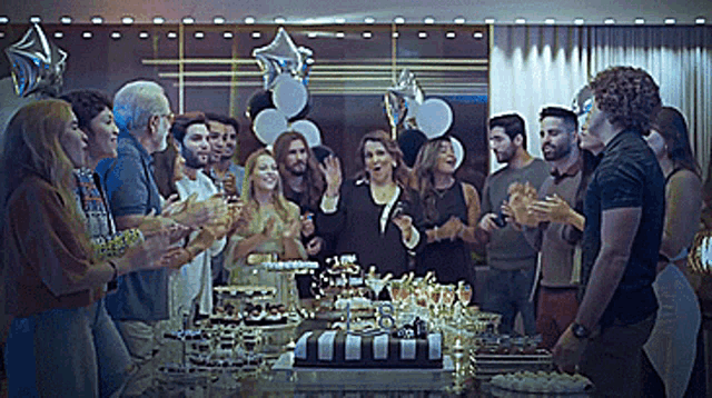 a group of people standing around a table with balloons and a cake