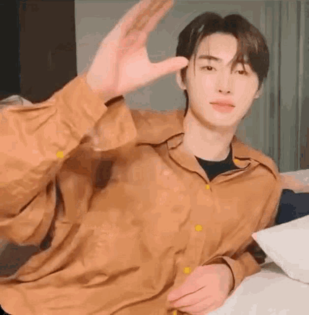 a young man in a brown shirt is sitting on a bed and making a peace sign with his hand .
