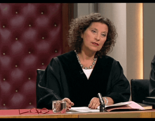 a woman in a judge 's robe sits at a desk with a book and a glass of water