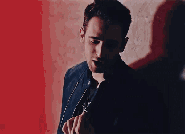 a man in a black jacket is standing in front of a red wall and looking at the camera .