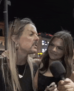 a man with long hair is talking into a microphone while a woman looks on