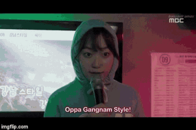 a girl in a hooded sweatshirt is holding a microphone and says oppa gangnam style
