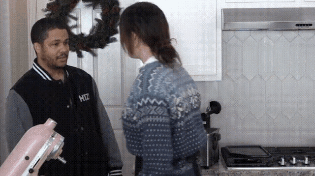 a man and a woman are standing in a kitchen and the man is wearing a jacket with the letter h on it