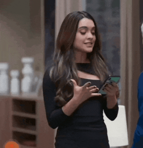a woman in a black dress is standing in a room holding a cell phone .