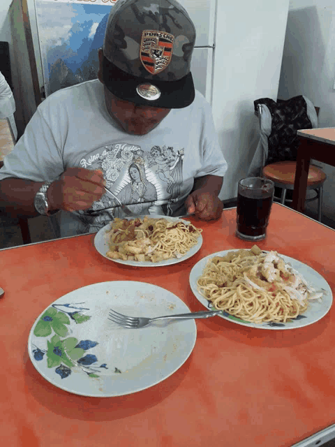 a man wearing a porsche hat is sitting at a table eating spaghetti