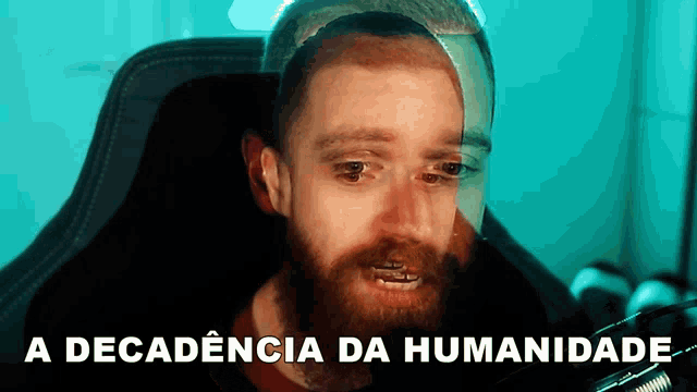 a man with a beard is sitting in front of a computer screen with the words " a decadencia da humanidade " behind him
