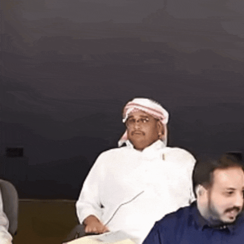 a man in a white shirt and a head scarf is sitting at a table with other people .