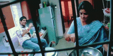 a woman in a blue saree is cooking while a man in a white shirt sits on a counter