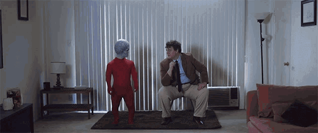 a man in a suit and tie sits on a couch next to a man in a red jumpsuit