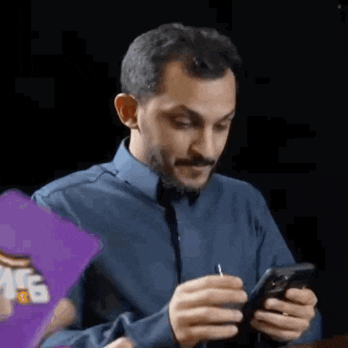 a man in a blue shirt and tie is sitting at a table looking at his cell phone .