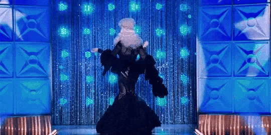 a drag queen is dancing on a stage with a blue curtain behind her .