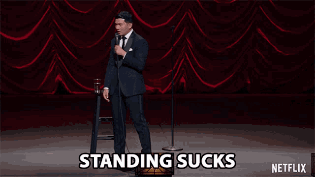 a man in a suit stands in front of a microphone with the words standing sucks written on the bottom