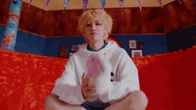 a young man in a white hoodie is sitting on a red couch holding a pink stuffed animal