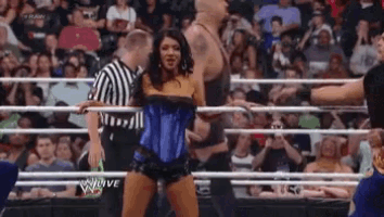 a woman in a blue corset is dancing in a wrestling ring while a referee watches .