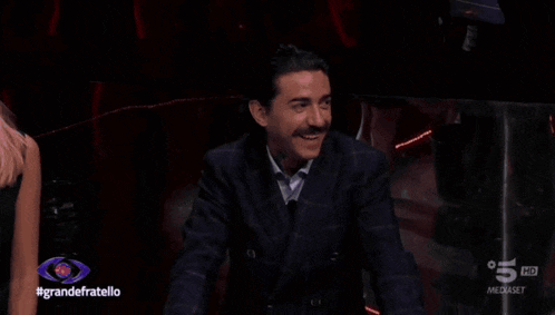 a man with a mustache is smiling in front of a sign that says #grandefratello