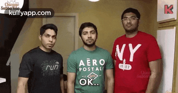 three young men are standing next to each other in a room . one of the men is wearing a red ny shirt .