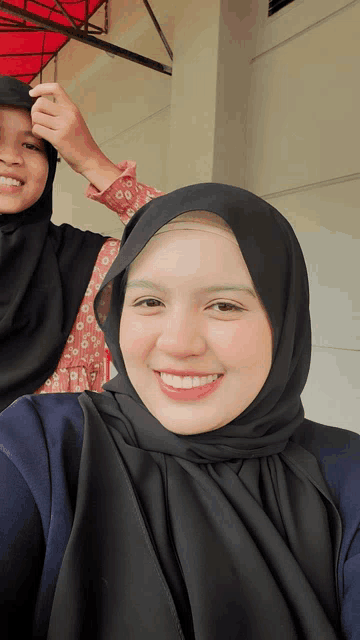 a woman wearing a black hijab smiles while another woman adjusts her hijab