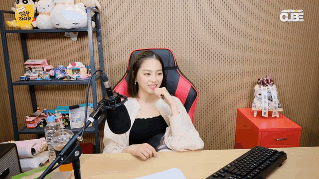 a woman sits at a desk in front of a microphone with a cube logo on the wall behind her