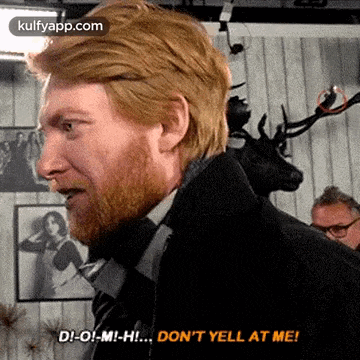 a man with red hair and a beard is standing in front of a wall with a deer head on it .