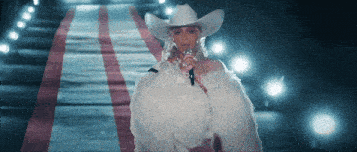 a woman wearing a cowboy hat and a fur coat sings into a microphone
