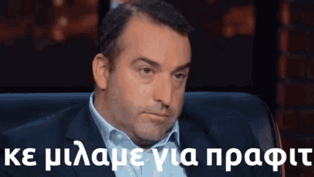 a man in a suit and blue shirt is sitting in a chair with a caption in greek