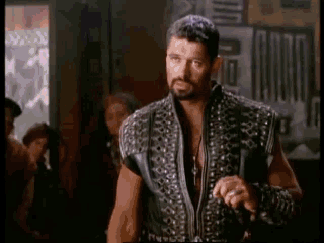 a man with a beard and a leather jacket is standing in a room in front of a group of people .