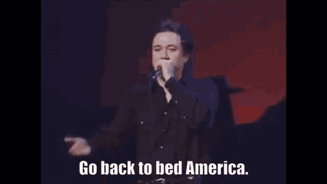 a man singing into a microphone with the words `` go back to bed america '' written below him .