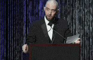 a bald man in a tuxedo stands at a podium with a microphone