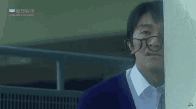 a man wearing glasses and a blue sweater is peeking out from behind a wall with chinese writing on it