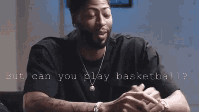 a man in a black shirt is sitting at a table with the words " but can you play basketball " behind him