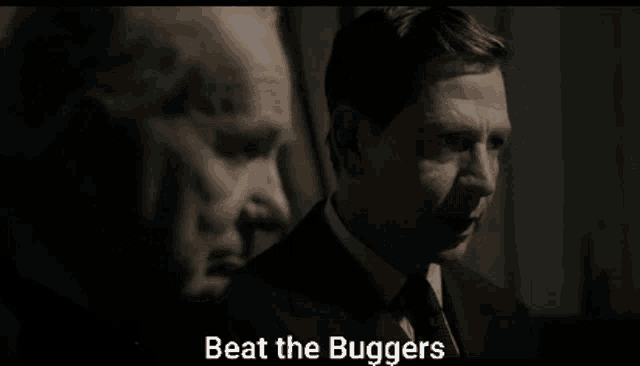 a man in a suit and tie is talking to another man in a dark room and the words beat the buggers are on the screen .