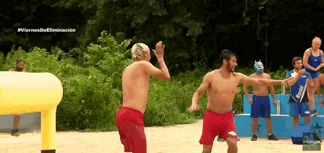 two shirtless men in red shorts are dancing on a sandy beach with the words #viernes de eliminacion on the bottom