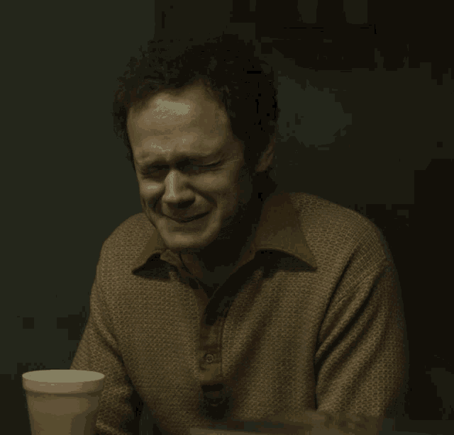 a man in a brown shirt is crying while sitting at a table next to a styrofoam cup