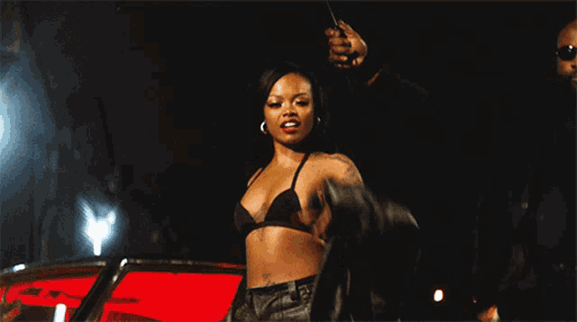 a woman in a black bra holds a gun in front of a red car