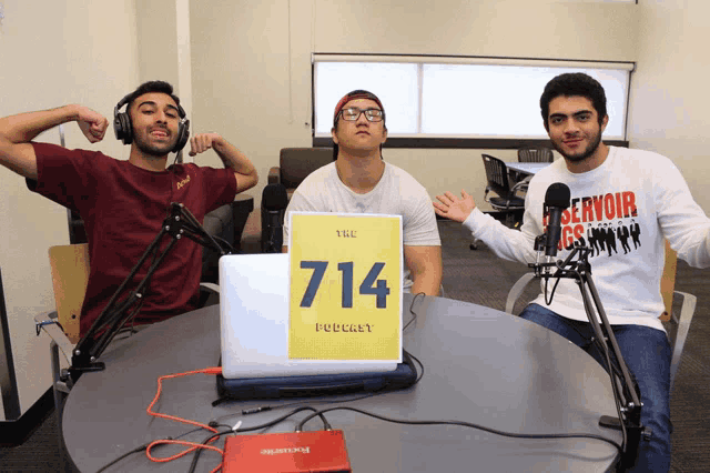 three men are sitting at a table with a laptop and a sign that says the podcast on it
