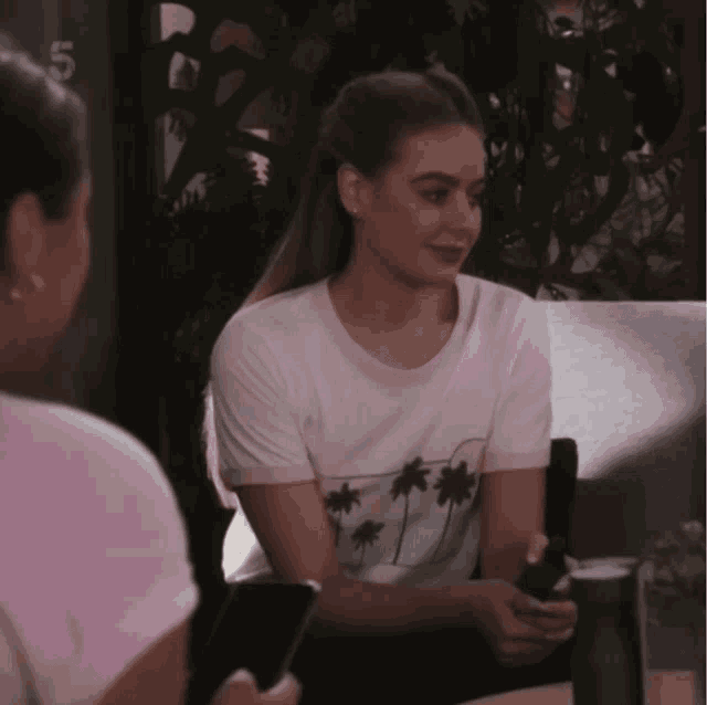 a woman wearing a white t-shirt with palm trees on it sits next to another woman