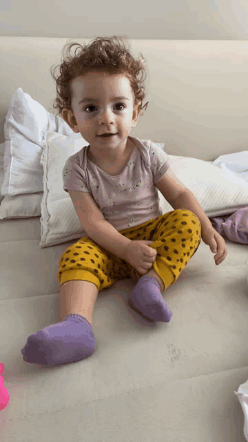 a little girl wearing purple socks and yellow pants sits on a bed