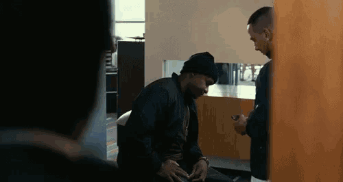 a man is sitting on the floor talking to another man while another man looks on .