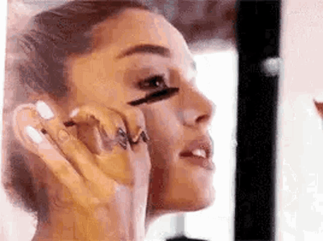 ariana grande is applying mascara to her eye while looking in the mirror .