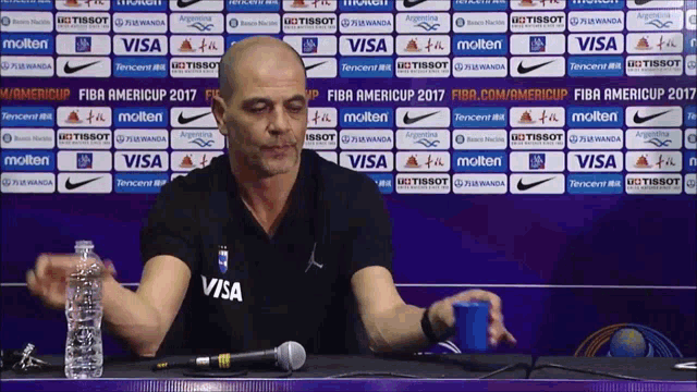 a man sitting at a table with a microphone and a bottle of water in front of a wall that says visa